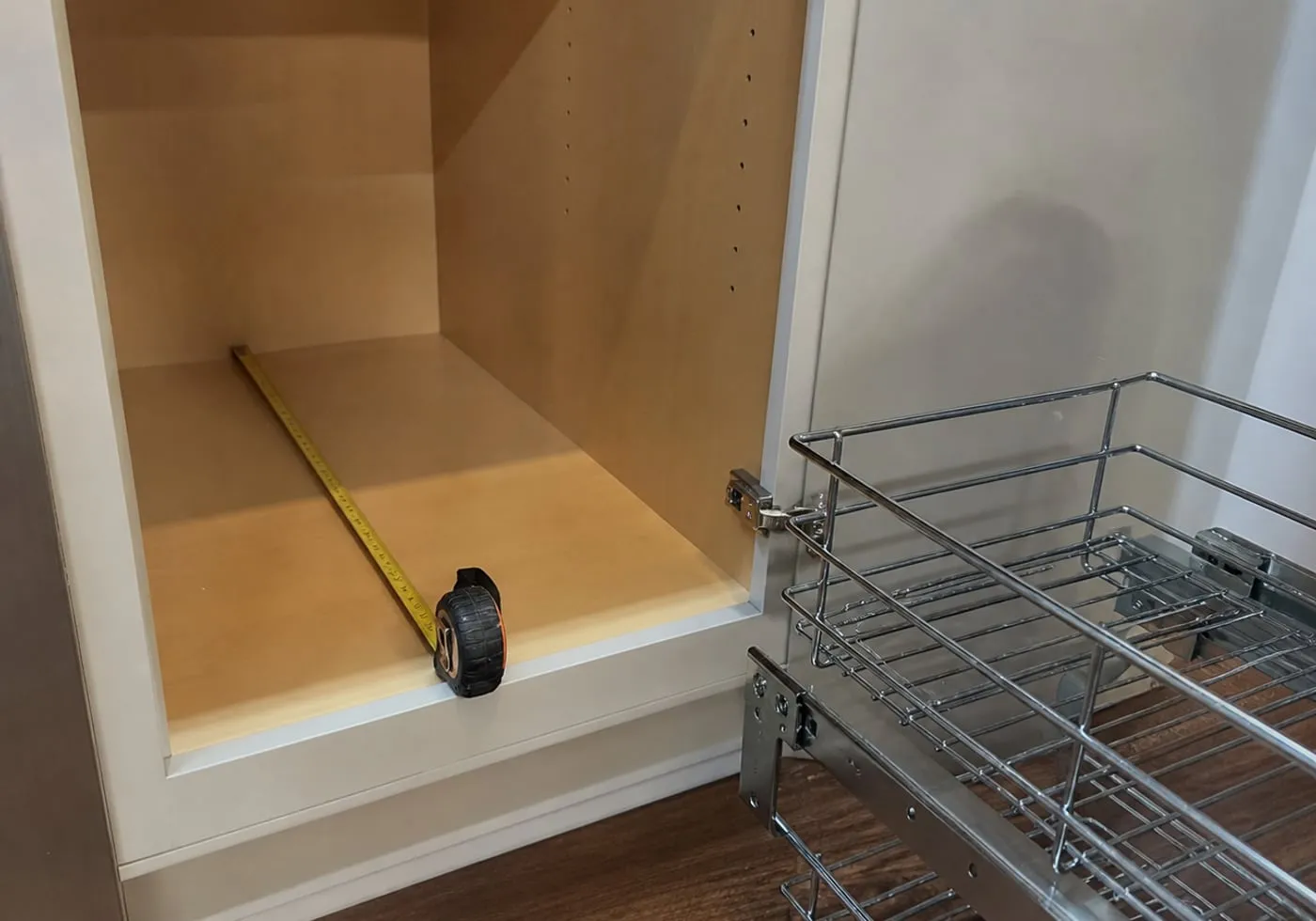 An empty lower kitchen cabinet being measured for inside depth, with a chrome wire sliding basket and rail set placed beside the opening