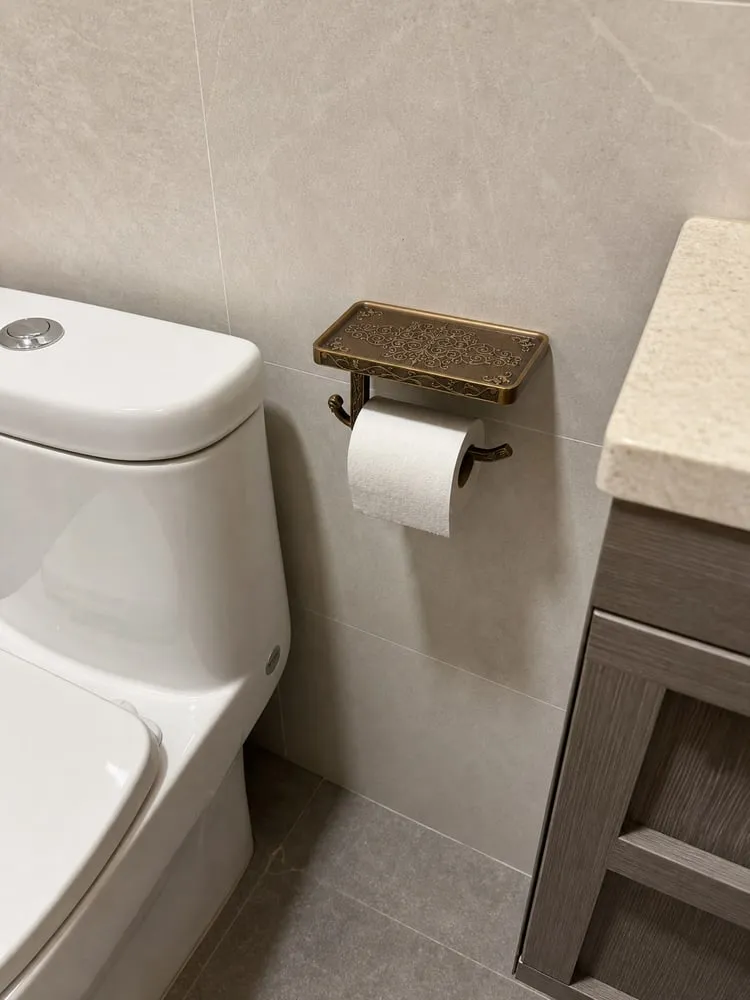 A side view of a shelf-style toilet paper holder near a toilet tank and bathroom cabinet edge