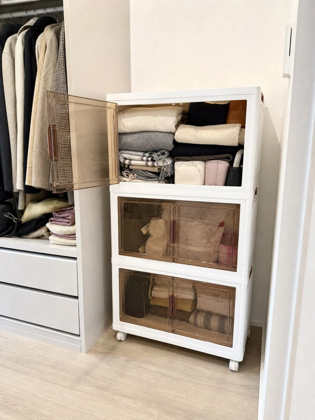 A white stackable storage bin inside a closet with one translucent front door open and folded clothes and small pouches inside