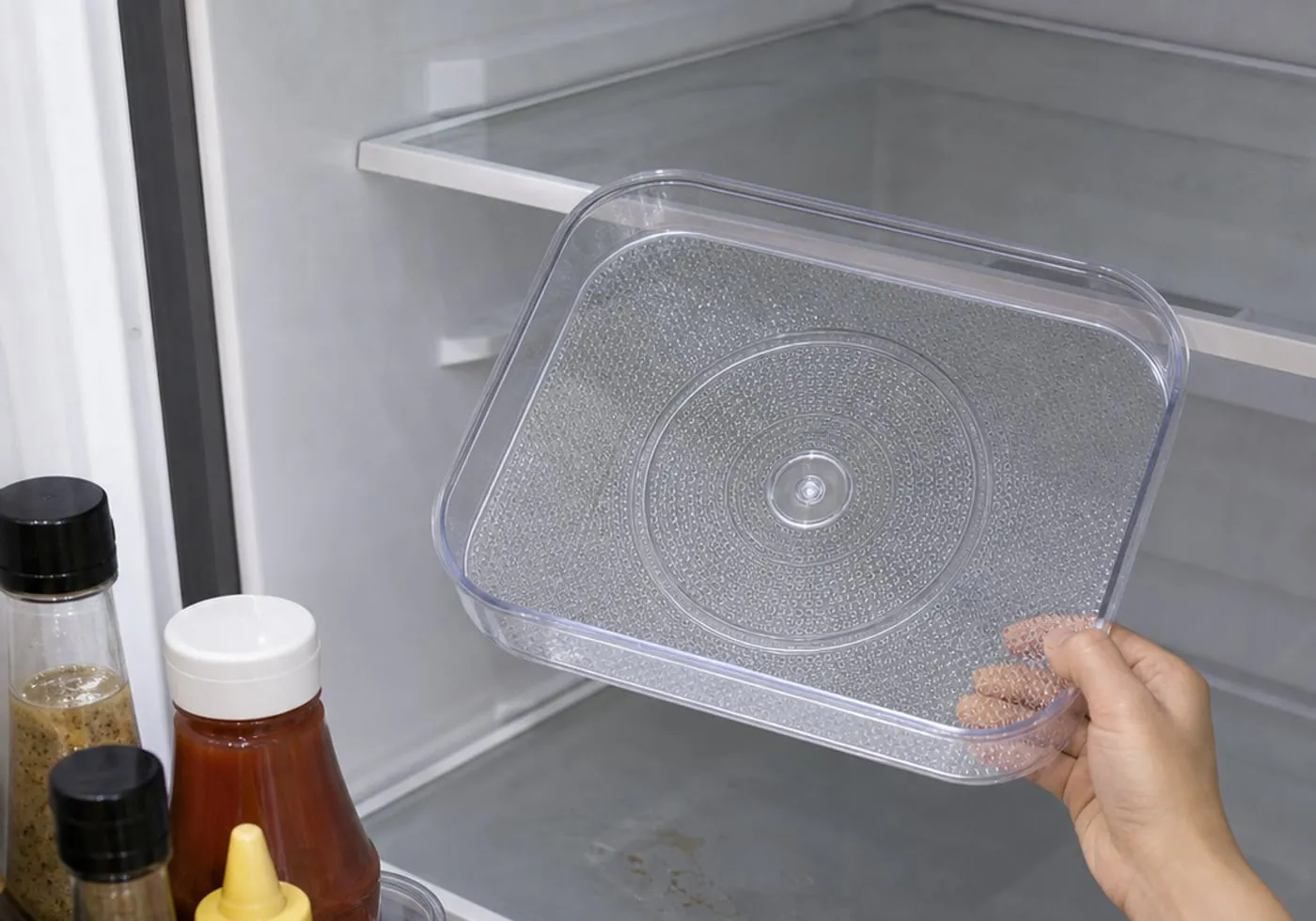 A clear fridge rotating tray lifted from the shelf while condensation and a small sticky spot are wiped underneath