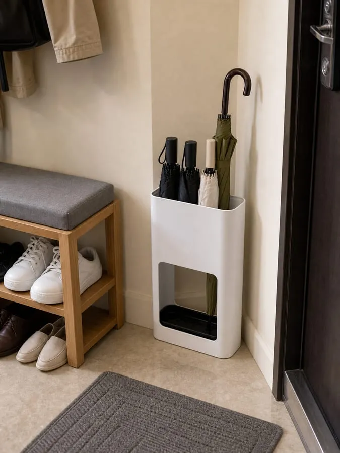 A white umbrella stand placed near an entryway corner with a modest number of umbrellas mostly upright and walking space left open