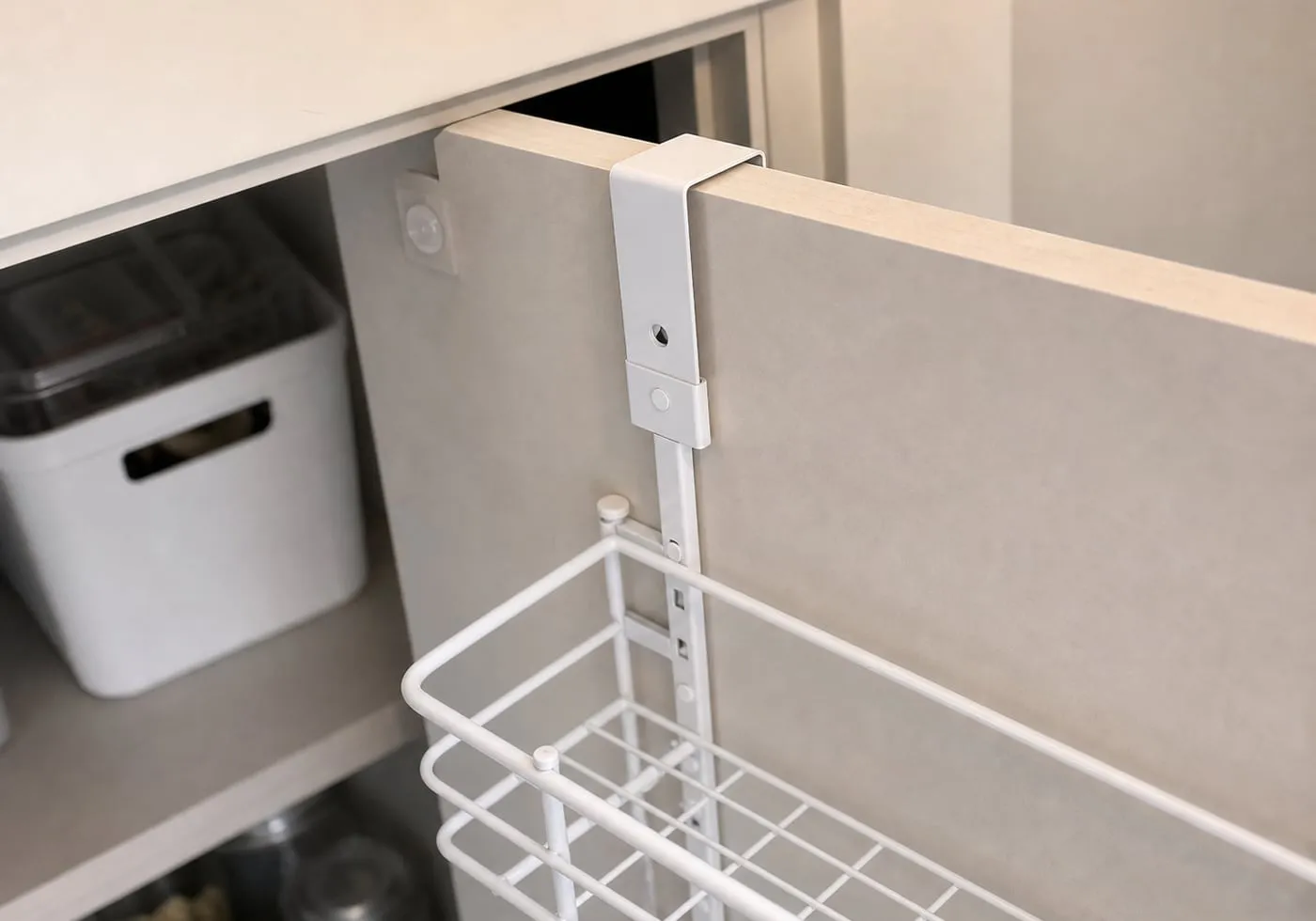 A close-up of a white metal over-door rack hook sitting over the top edge of a pantry door with a narrow frame gap and a small protective pad nearby