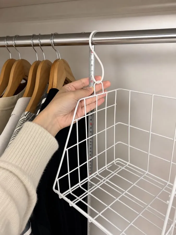 A hand checking the S hook of a white wire basket against a closet rod, with the empty wire basket and its depth visible
