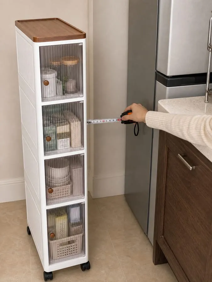 A hand measuring the narrow gap between a refrigerator and kitchen cabinet while the white slim rolling cabinet waits nearby