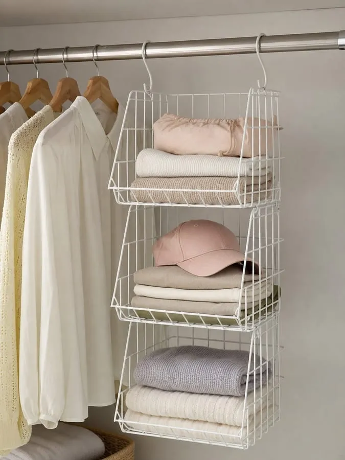A white wire hanging basket organizer under a closet rod with folded cloths, a hat, and a small pouch separated into light layers