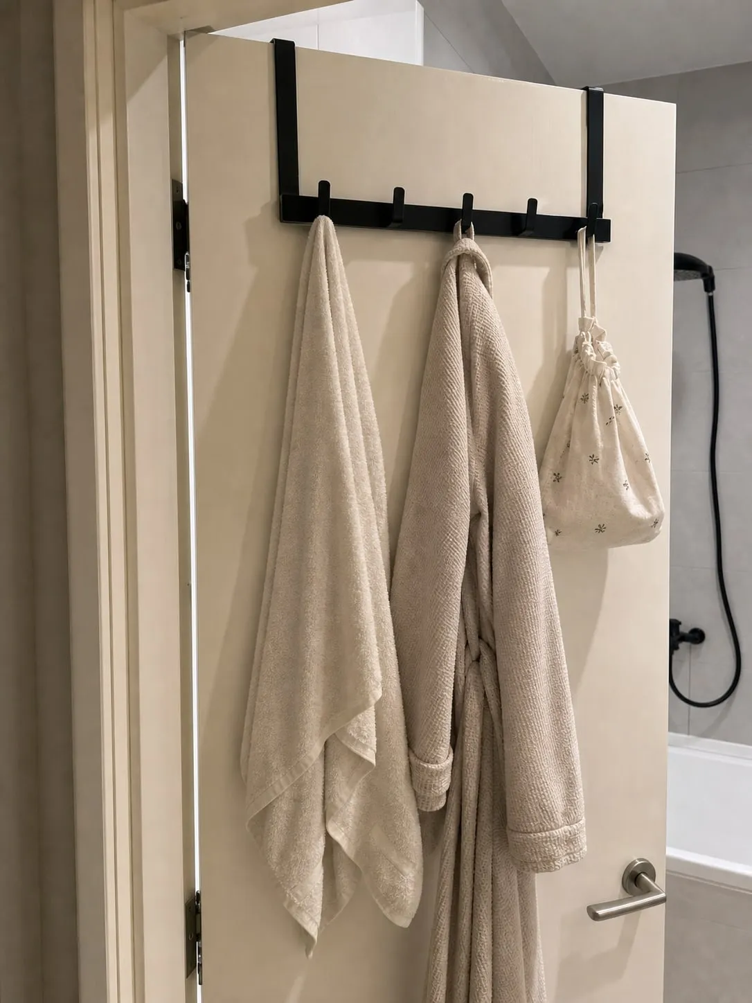 A black over-door five-hook rack on a white bathroom door with a towel, robe, and small fabric bag hanging separately