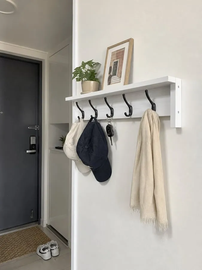 A white wall-mounted shelf rack near an entryway with black hooks holding caps, keys, and a light scarf