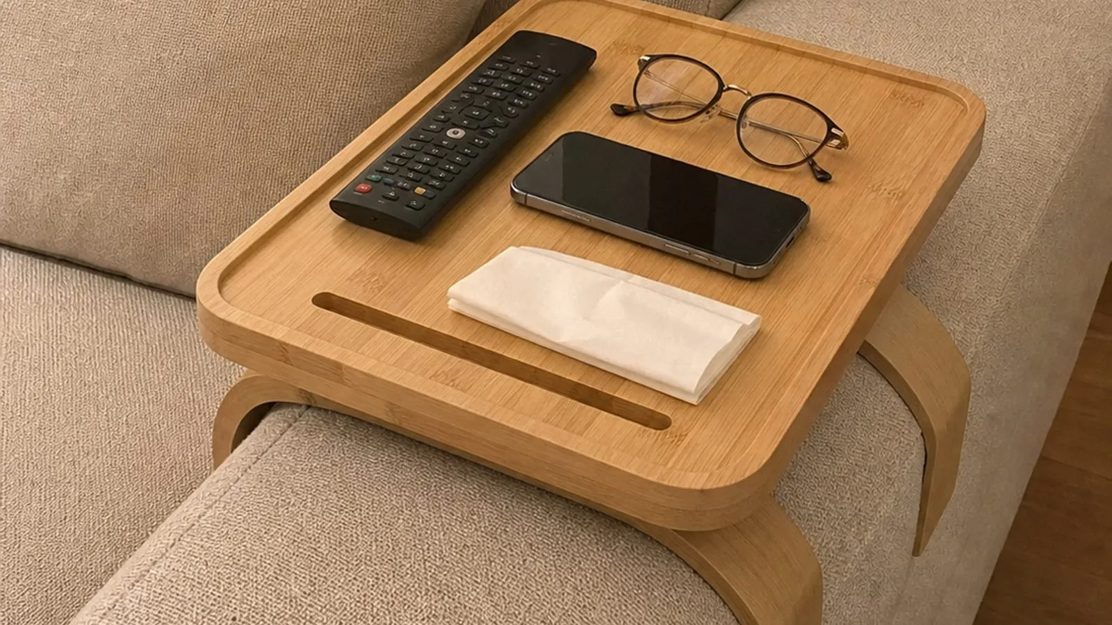 Light wood sofa-arm tray on a wide fabric sofa arm with a remote, phone, glasses, and tissue
