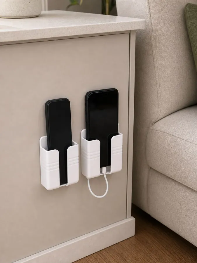 Two small white wall-mounted plastic holders installed low beside a sofa, holding a remote turned inward, one phone, and a short loose cable