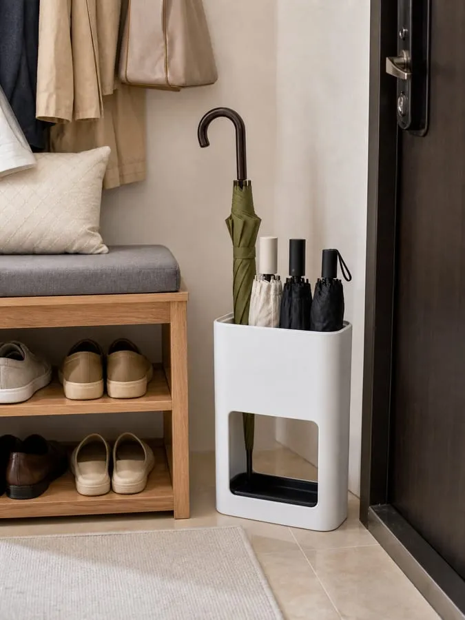 A white rectangular umbrella stand beside an entryway shoe bench, holding one long umbrella and several folding umbrellas with a black lower tray visible
