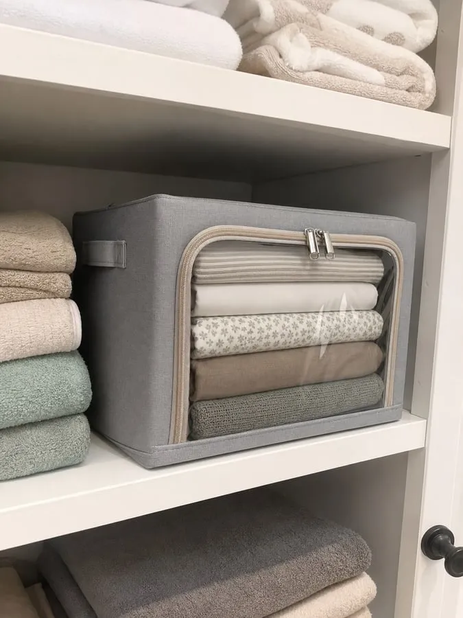 A gray framed fabric bedding storage box on a linen closet shelf with folded sheets and a thin blanket visible through the clear front window