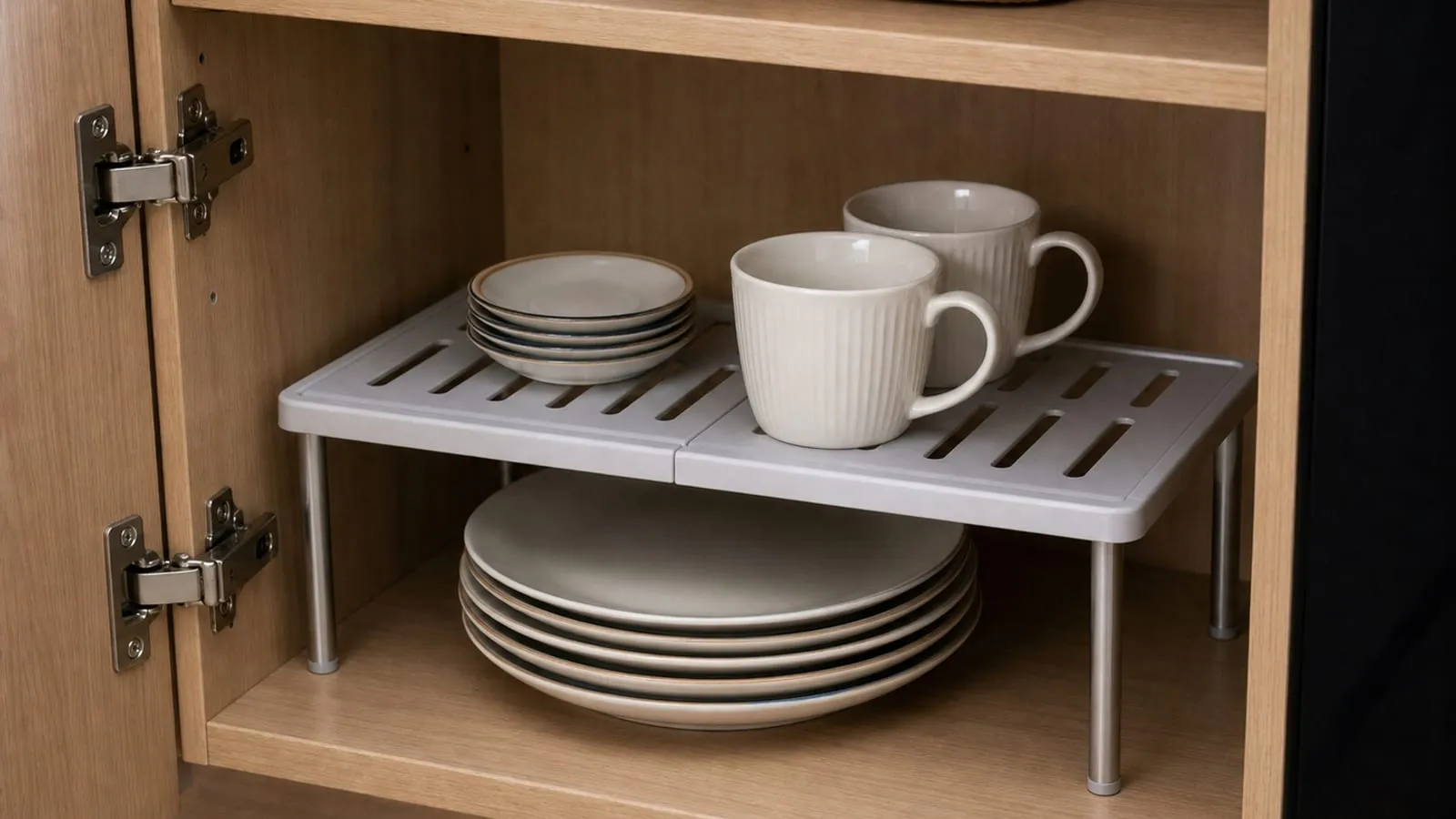 Gray slotted cabinet shelf riser inside a kitchen cabinet with plates below and cups on the upper level