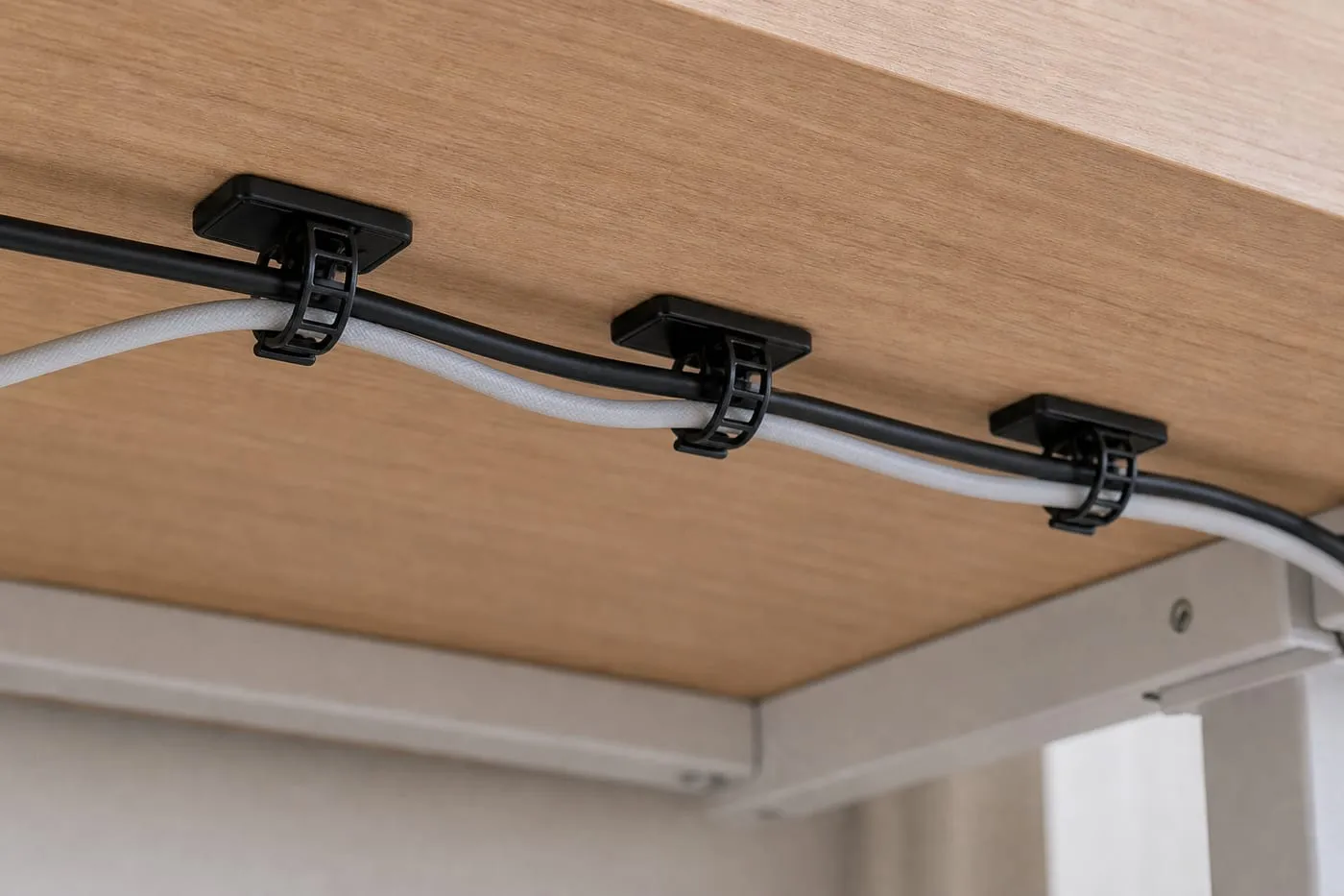 Black strap-style adhesive cable clips under a desk guiding light cables with slack, without holding any adapter weight