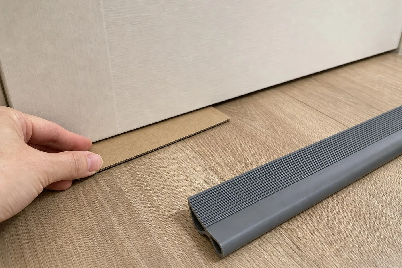 A hand checking the under-door gap with an unmarked cardboard spacer while a grey door-bottom strip sits nearby