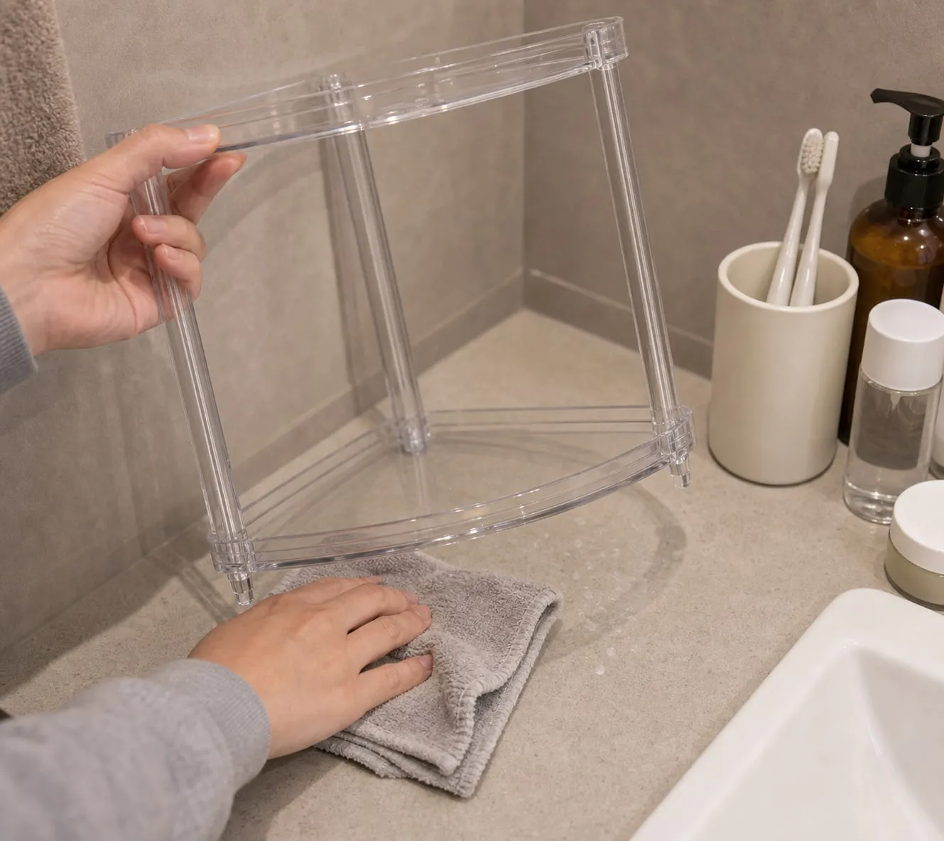 A clear acrylic bathroom corner shelf being lifted slightly while water spots underneath are wiped with a cloth