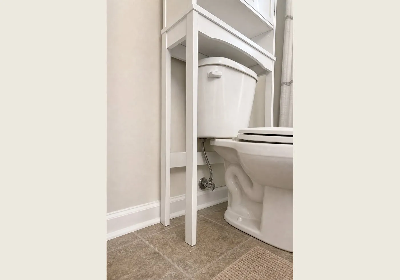A low side angle showing a white over-toilet cabinet leg placed near toilet plumbing and hose clearance without touching them