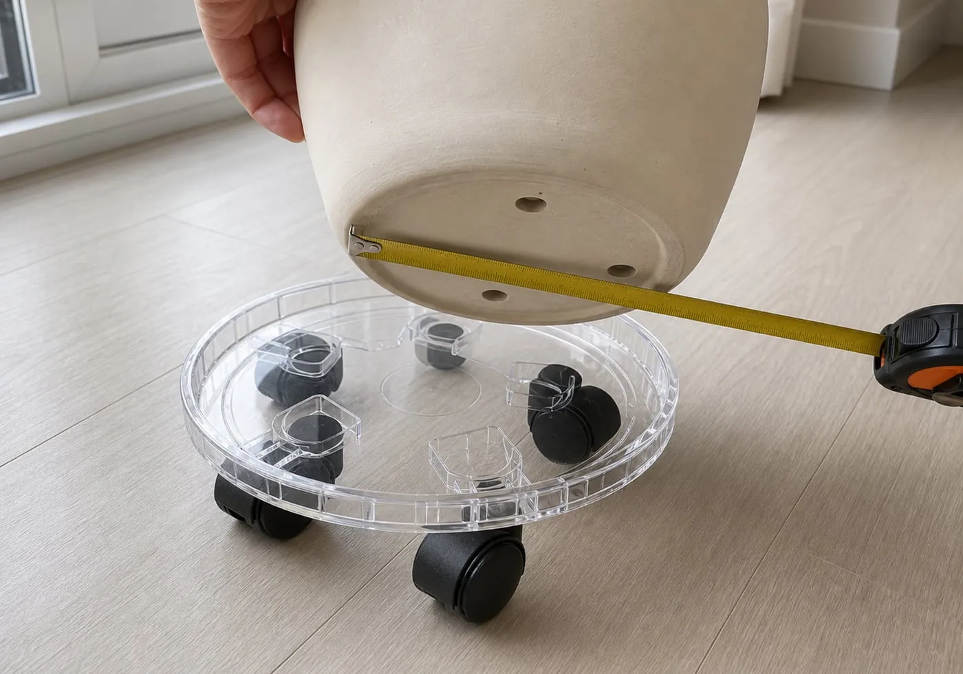 A close-up of a pot bottom being checked against a transparent round rolling plant caddy with a tape measure and black caster wheels visible