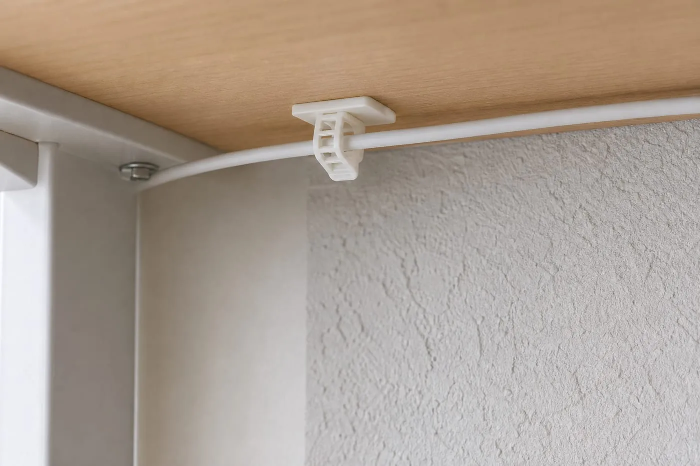 A white strap-style adhesive cable clip attached to a smooth desk underside while a nearby textured wall area remains unused